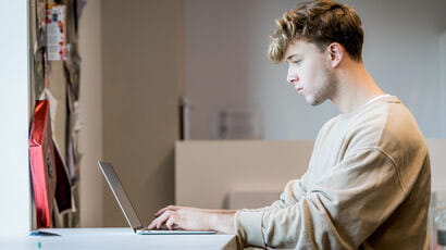 Two students are sitting in student accommodation, using a laptop and a textbook.