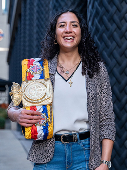 Nicola, BSc(Hons) Biomedical Sciences smiling to camera with a huge boxing medal belt hanging over her shoulder.