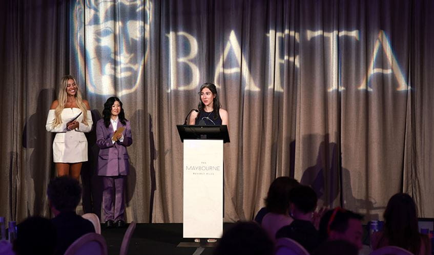 A woman stands at a lectern making an acceptance speech for an award, with the BAFTA logo behind her.