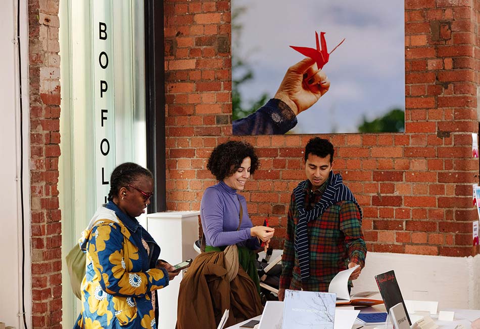 Three global majority employees standing at a desk reviewing books in an office with photography hung on exposed brick walls.