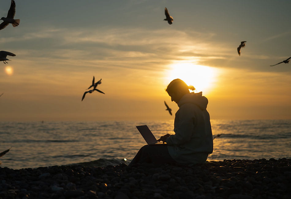 A person sitting at the beach at sunset with lots of birds flying in the sky above.