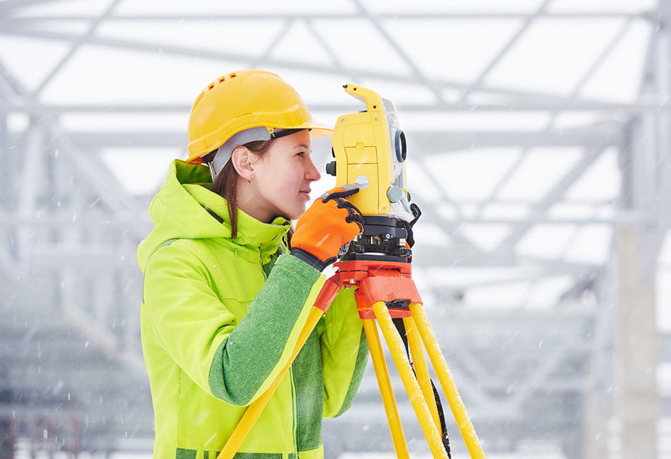 A student wearing a helmet and reflective jacket while using surveying equipment.