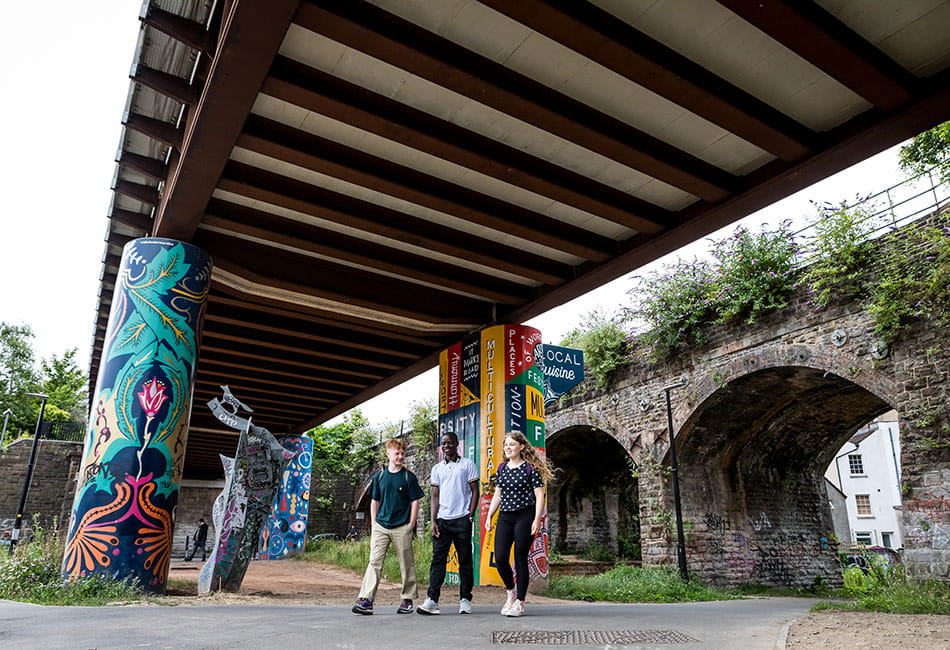 Three people walking under a road bridge with colourful grafiti in a suburban area.