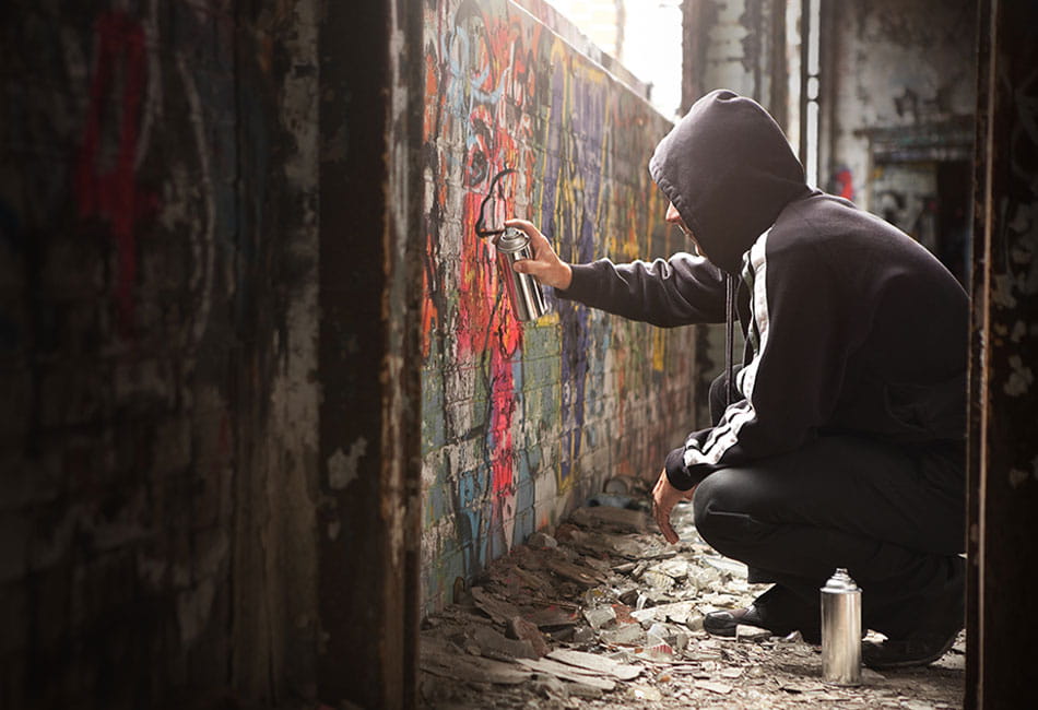 A man wearing a black hoodie spraying graffiti on a wall in an abandoned house.