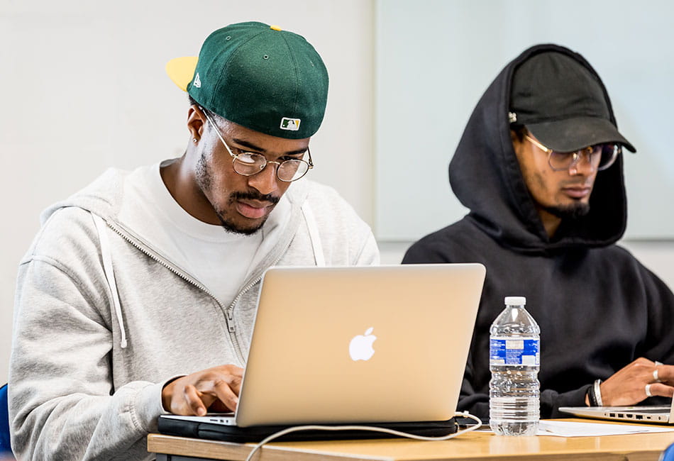 Two students wearing hoodies and caps studying on laptops in a classroom.