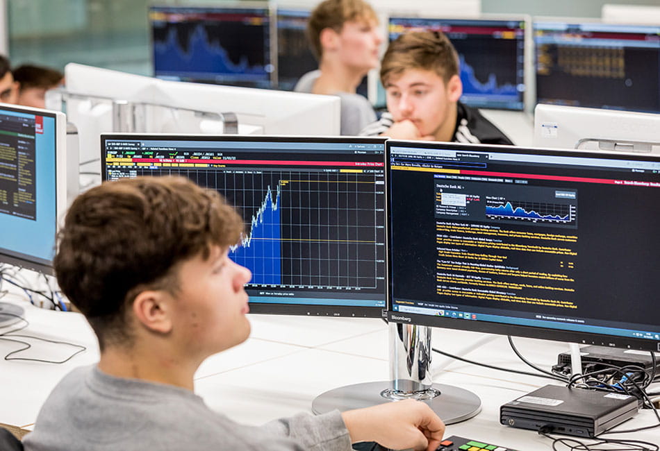 Students studying charts and graphs on double monitors in a Bloomberg trading room.