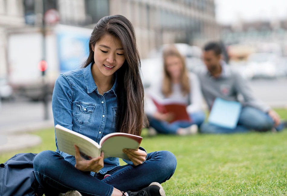 A student reading a book seated cross-legged on the grass.