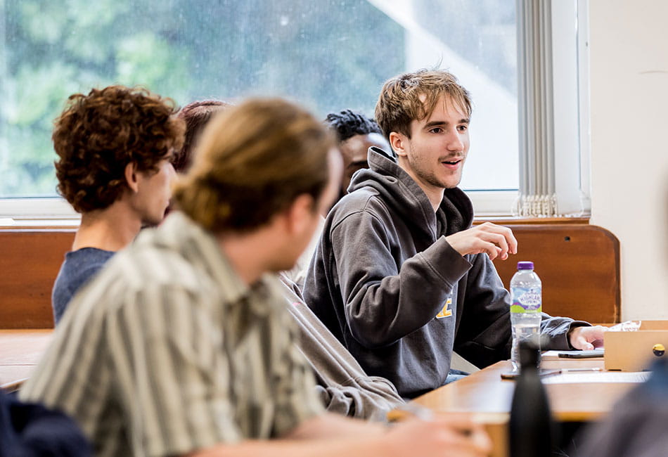 A group of students sat in a classroom listening whilst one of them talks.