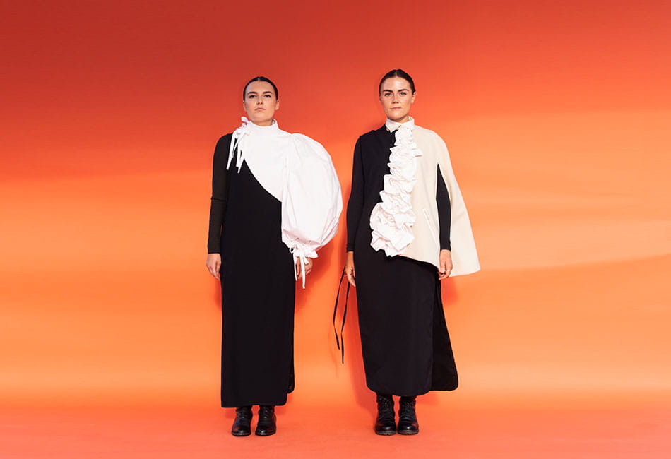 Two models wearing black and white dresses standing in front of an orange background.