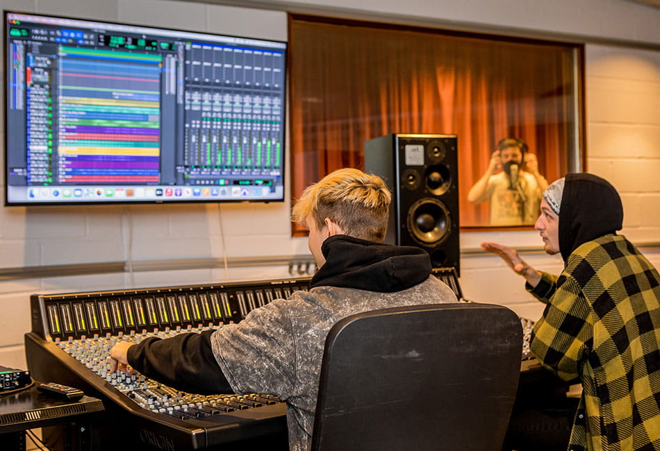 Two students using the audio workstation equipment whilst a student sings in the recording studio.