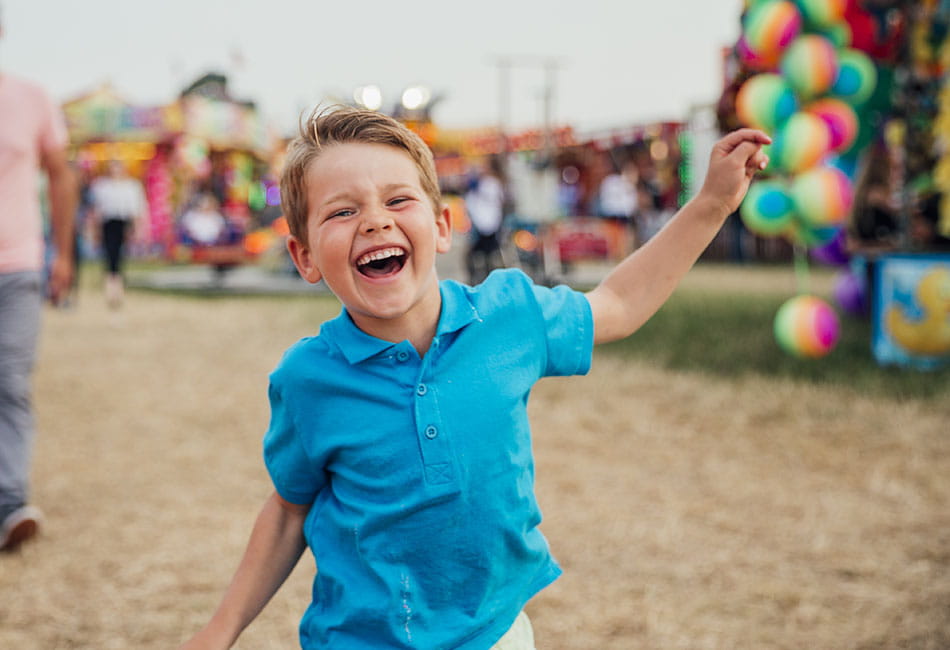A smiling boy in blue polo shirt running around a fair.