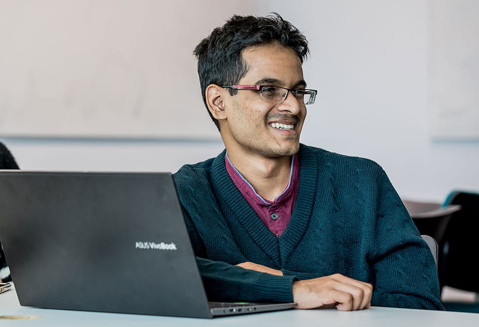 A student smiling to a friend off camera while at his laptop in a study space.