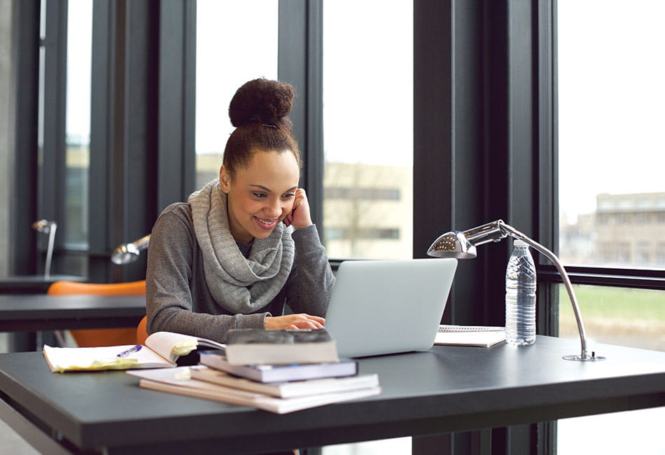 A student sat at a desk looking at a laptop with books surrounding her.