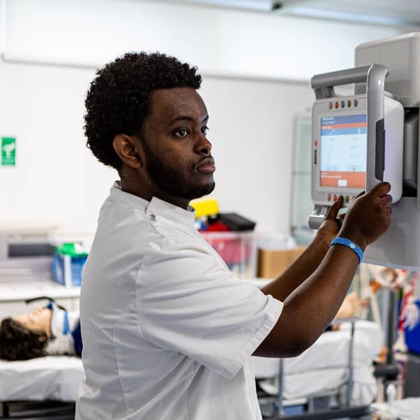 A black man wearing a white medical outfit has his hands placed on a diagnostic machine. There is a dummy patient on a bed in the background.