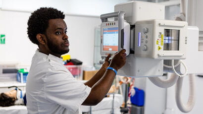 A black man in a white medical practitioner outfit with his hands on a diagnostic machine with a screen on it. A dummy patient lies on a bed in the background.