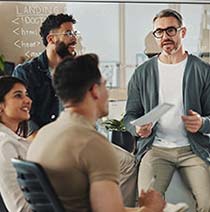 A modern relaxed office meeting room where a group sit closely in front of a whiteboard with a man presenting.