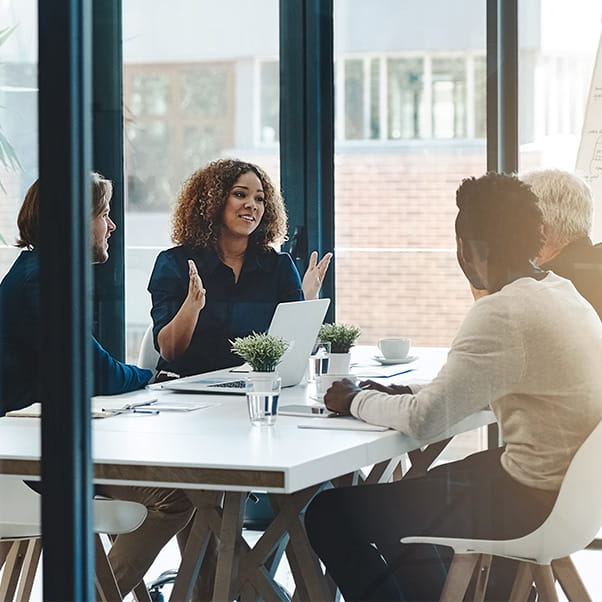 A view into a glass walled office where four co-workers in smart dress sit together around a meeting table.