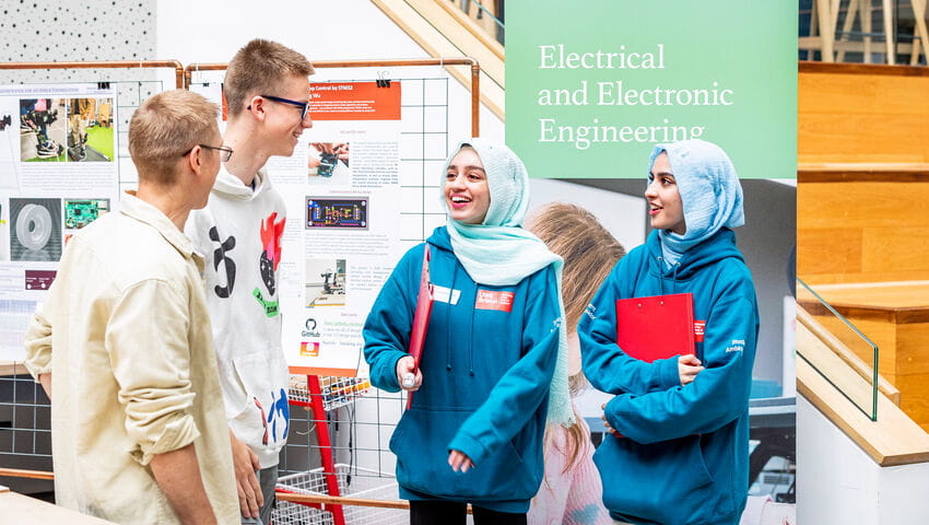 Two Student Ambassadors talking to a prosective student and their family member. A banner that says Electrical and Electronic Engineering stands in the background.