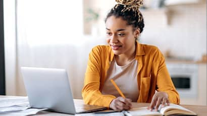 Young adult watching a webinar on their laptop at home and taking notes.