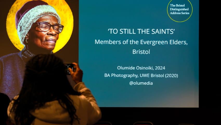 Projector on stage at a BDAS event with photography of a black woman with her eyes closed in front of a golden moon and accompanying text that says 'TO STILL THE SAINTS'.