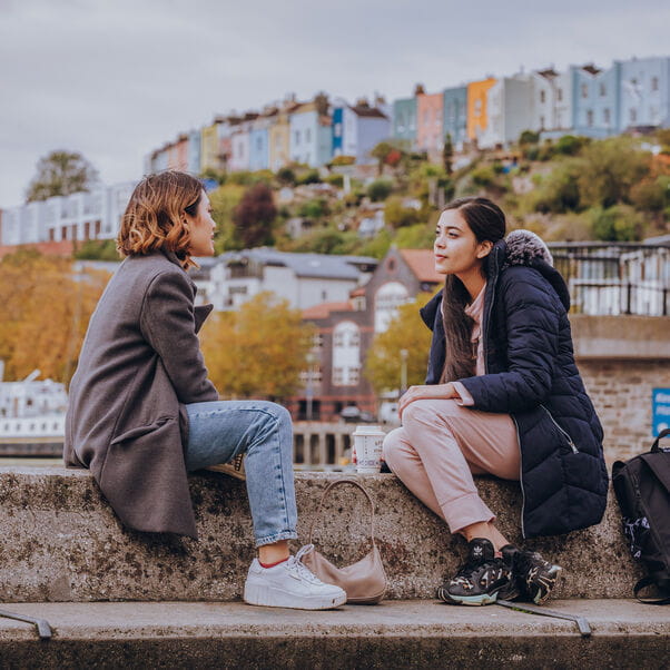 Two students sitting on a wall by the harbourside talking to one another.