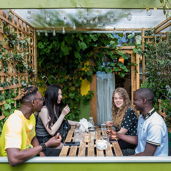 Group of four students sat together at a wooden table and bench in a covered outdoor space covered with light bulbs and greenery.