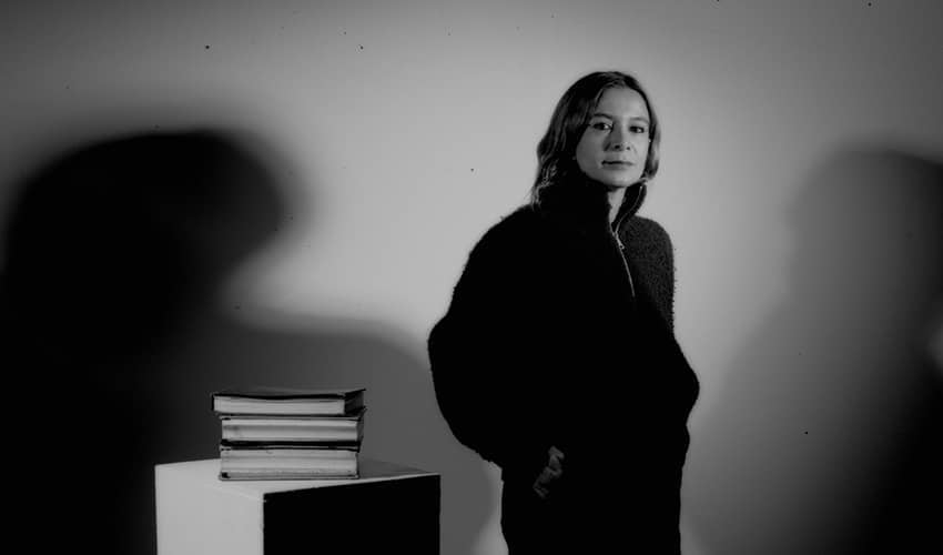 Black-and-white photograph showing a person standing in front of a plain wall with strong shadows cast on both sides. To the left, there is a small pedestal or table with a stack of books placed on top.