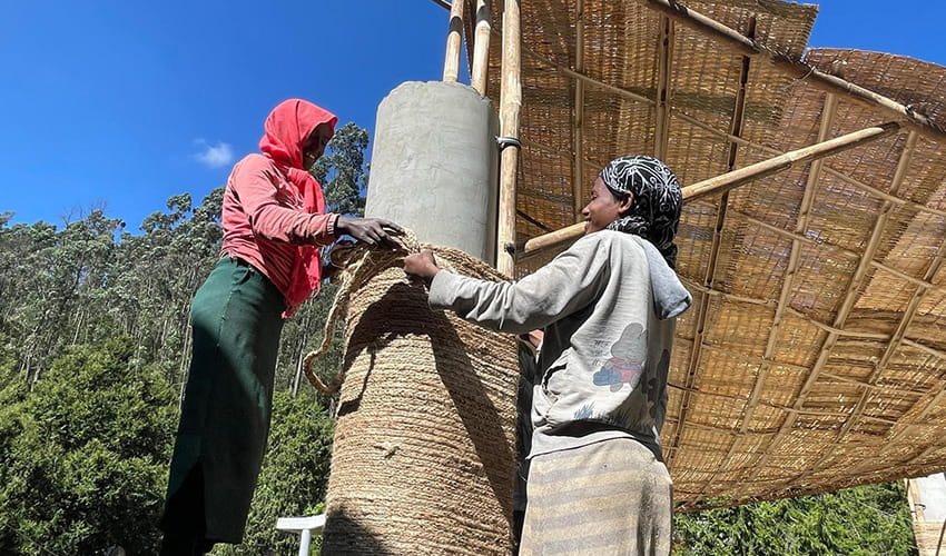 Two people covering a large pillar in brown rope as part of a large pavilion structure 