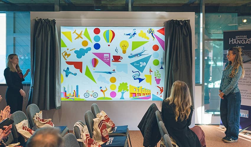 A colourful, abstract collage featuring various transportation and lifestyle elements is unveiled in front of a group of people.