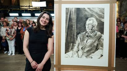 A woman wearing a black outfit stands beside a large portrait, with a crowd of people gathered in the background