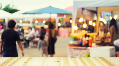 An outdoor event with food vendors surrounding some tables and chairs with people sat at them.