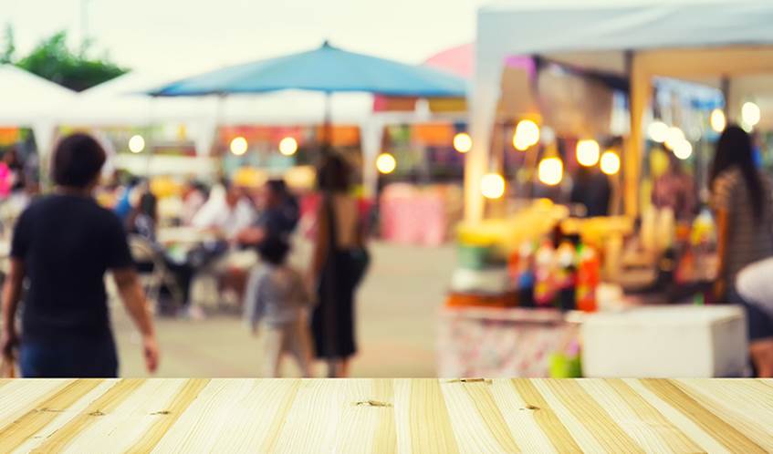 An outdoor event with food vendors surrounding some tables and chairs with people sat at them.