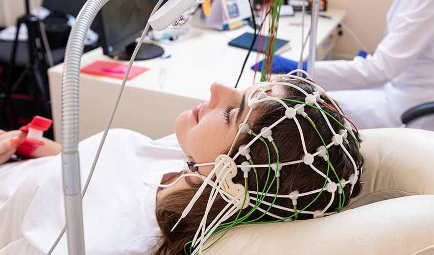 A woman is lying down in a hospital with a series of wires covering her head to monitor brain activity.