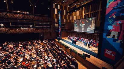 A large auditorium filled with an audience watching a stage with a graduation ceremony taking place and a student in a cap and gown walking across the stage.