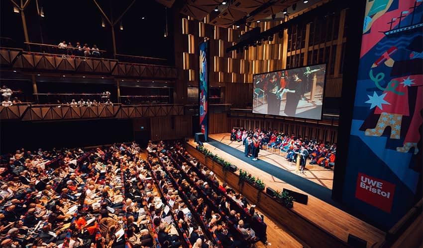 A large auditorium filled with an audience watching a stage with a graduation ceremony taking place and a student in a cap and gown walking across the stage.