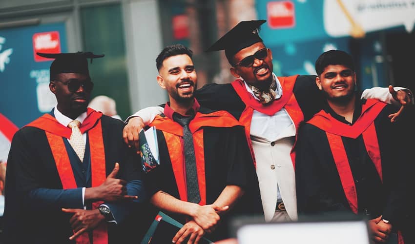 A group of four graduates wearing black caps and gowns with red stoles stand closely together, posing for a photo.