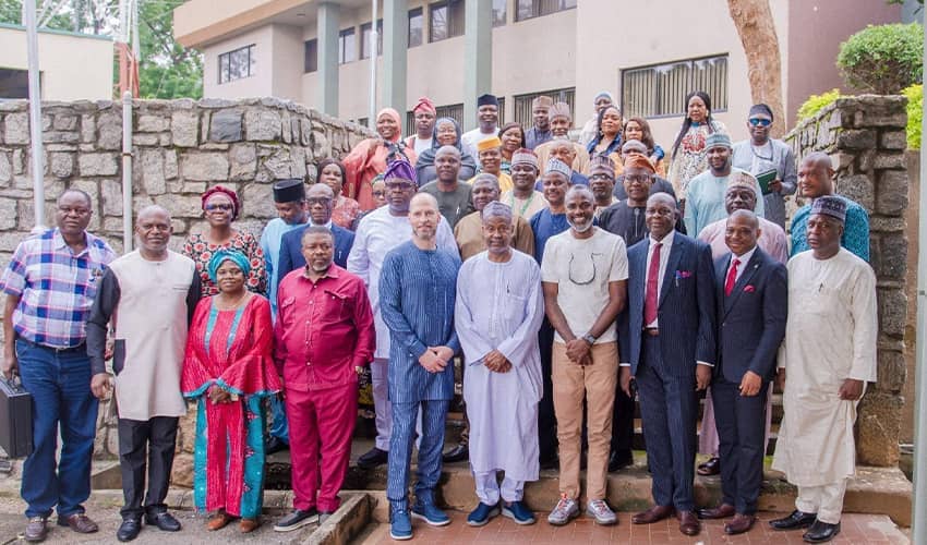 A large group of people standing on steps in front of a stone wall and buildings, posing for a photo. The group is dressed in a mix of traditional African clothing and suits. 