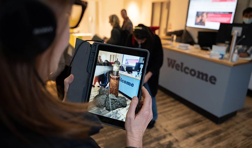 A woman stands in an office looking at a mobile phone screen which shows a virtual reality image of a lighthouse standing on rocks.