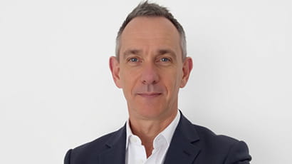 A man wearing a dark suit and white shirt looks at the camera.