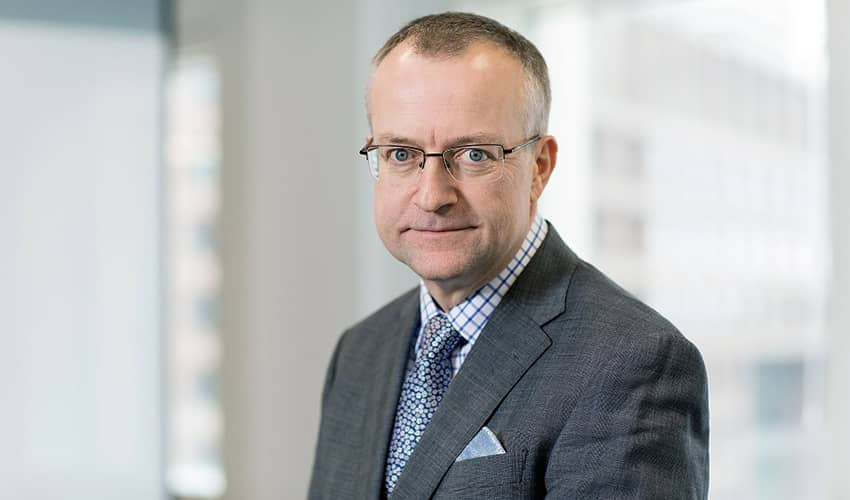 A head and shoulders image of a man wearing a suit with glasses looking down the lens of the camera in a corporate setting. 