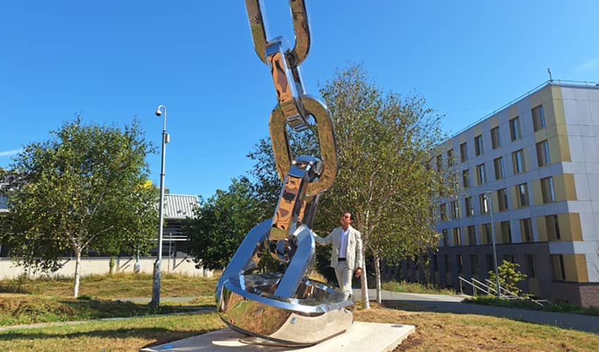 A large stainless steel structure of multiple links connecting outside a university campus with a man stood resting on the sculpture looking up at it