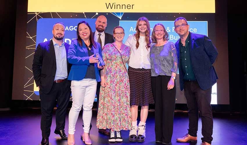 Seven people in a group smiling at the camera at an awards ceremony, with the word 'winner' displayed on a screen above their heads