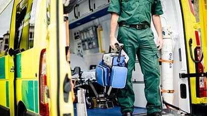 Male paramedic standing in the back of an open ambulance, carrying a bag of kit in one hand