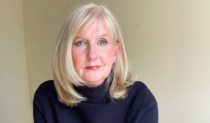 A woman with blonde hair and a black jumper looking at the camera against a beige background