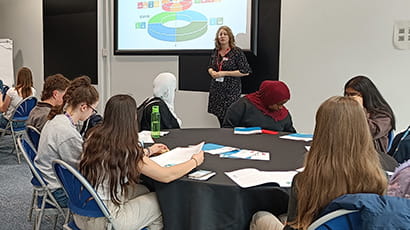 A classroom, with a lecturer stood in front of a white digital screen in the background, and secondary school students sat at a round table in the foreground