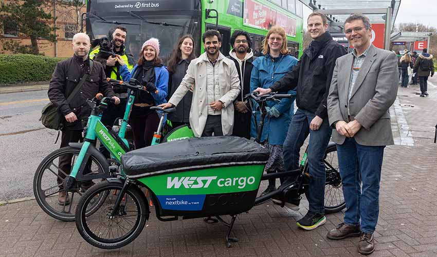 A  group of nine people stood behind a green cargo bike and a green electric bike, with a bus at a bus stop in the background