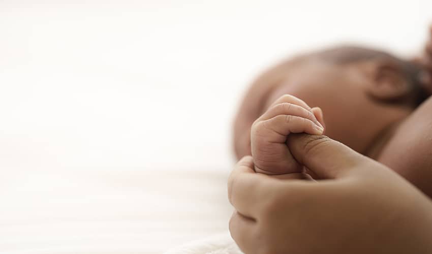 Image of a newborn baby, with side of its face blurred in the background, and a parents hand holding the baby's hand in the foreground