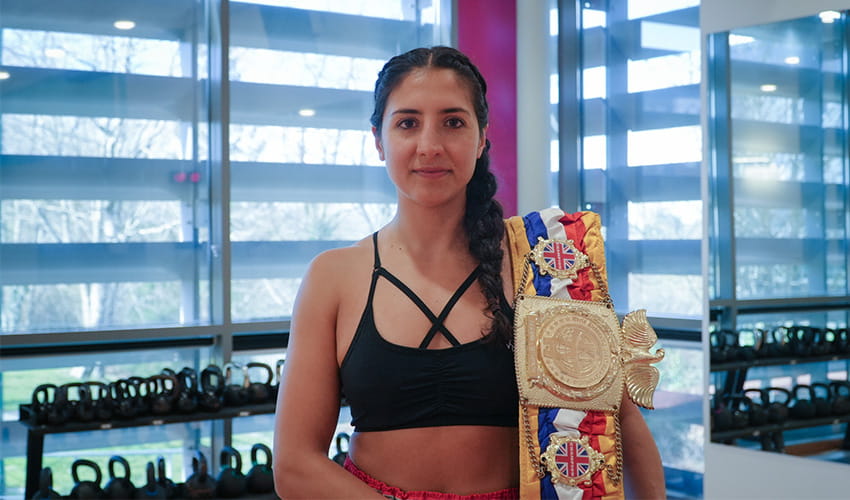 A woman in a gym with a martial arts championship winner belt over her shoulder 