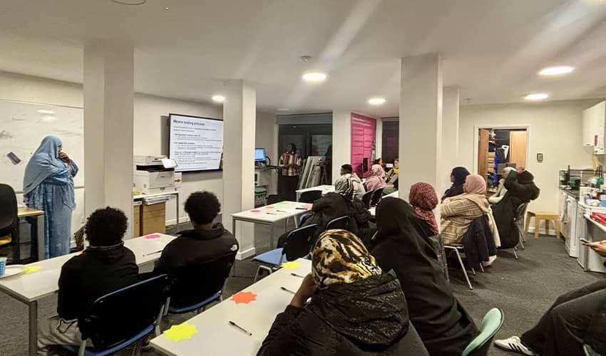 A classroom setting with two rows of tables with people sat at them facing a person stood at the front of talking to the group.