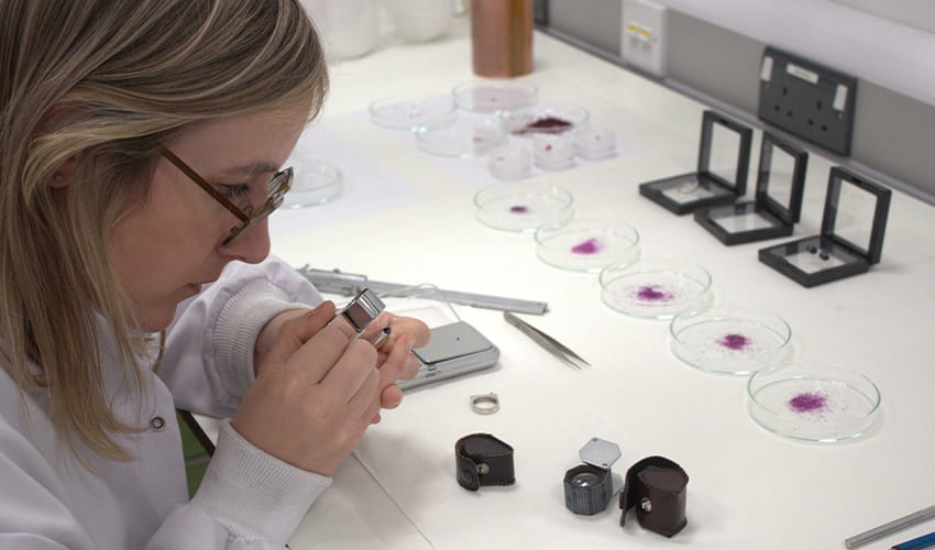 A woman working in a lab examines a ruby gemstone with a magnifying glass